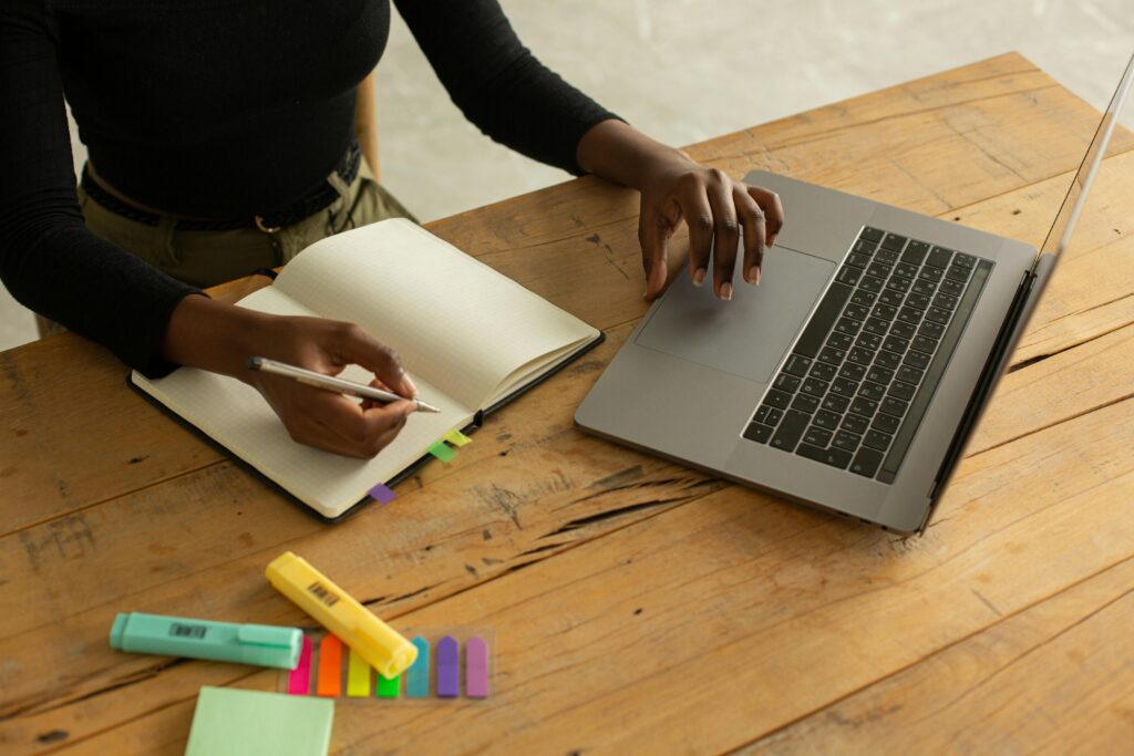 Person taking notes on laptop and notebook with a pen, highlighters, and a notepads.
